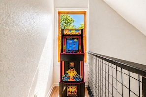 Una máquina arcade de Pac-Man en un pasillo con una escalera y un ventanal con vistas a árboles.