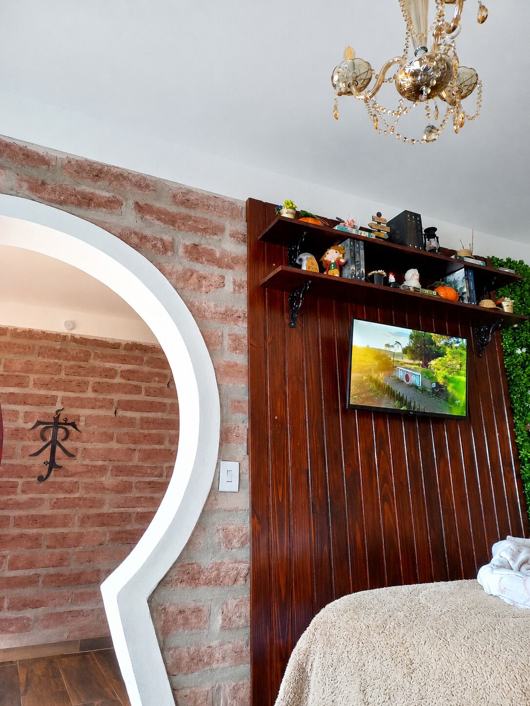 A room with a brick wall, a wooden shelf with various items, a TV, and a chandelier.