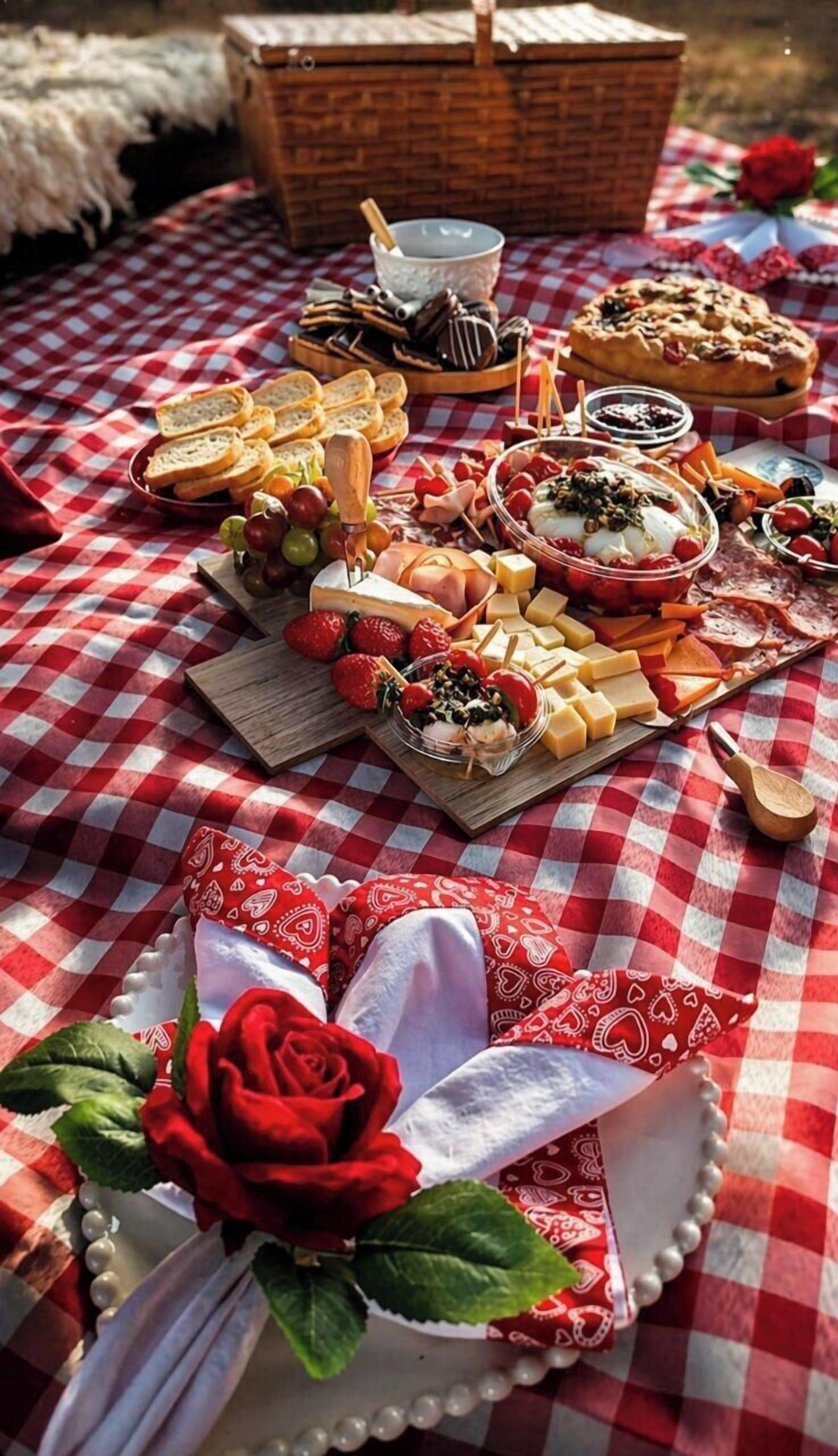Een picknickopstelling met een geruit tafelkleed, diverse kazen, fruit en een rode roos.