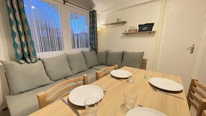 Un coin repas avec une table en bois dressée pour quatre personnes, un canapé avec des coussins et une fenêtre ornée de rideaux légers.
