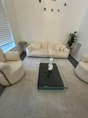 Ein Wohnzimmer mit einem beigen Sofa, zwei weißen Sesseln, einem Glastisch mit einer weißen Vase und einer Wanduhr.