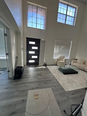 Ein geräumiges Wohnzimmer mit hohen Decken, großen Fenstern, einem modernen Sofa, einem Glastisch und einem gemusterten Teppich.