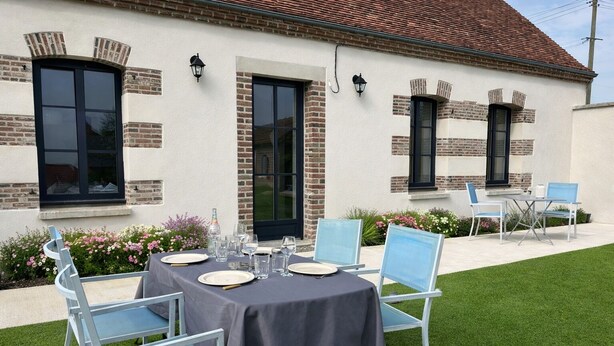 An outdoor dining area with a table set for four, surrounded by light blue chairs, against a building with brick accents and large windows. - Charming, Renovated Farmhouse • Private garden • Terrace • Calm • Up to 6 people. (saviere)