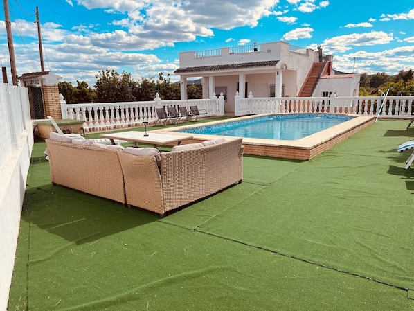Une piscine sur le toit, entourée d’une clôture blanche, avec un canapé en osier et un immeuble de deux étages en arrière-plan.