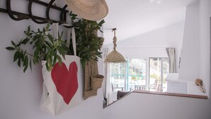 Quarto com parede branca, um porta-chapéus pendurado, um vaso de planta e uma luminária suspensa, com vista para o exterior pela janela.