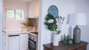 Cozinha com armários brancos, máquina de lavar louça, forno, janela com persianas azuis, um vaso com flores, um prato decorativo e uma luminária.