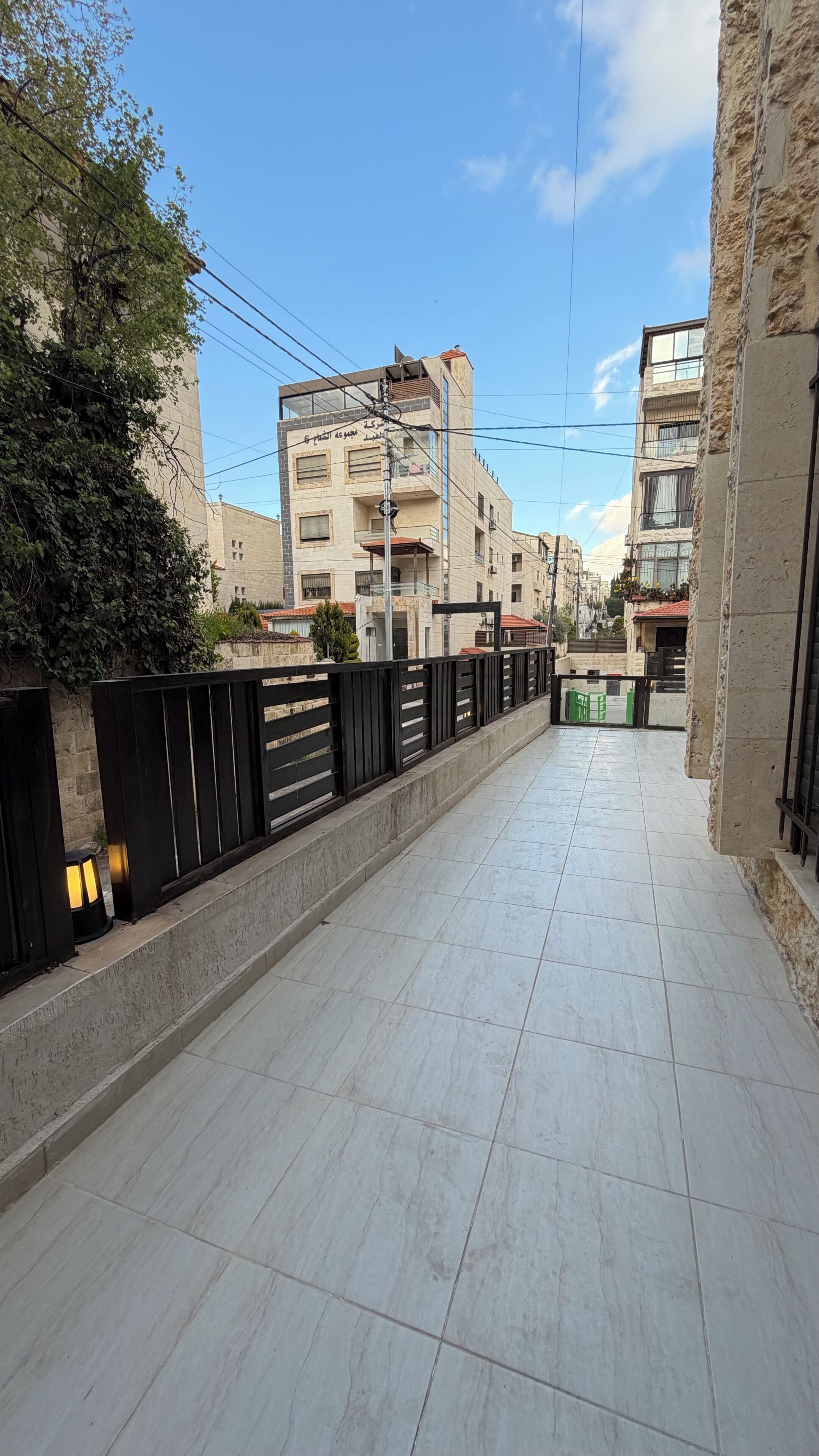 A corridor with tiled flooring, a black metal fence, and buildings in the background.