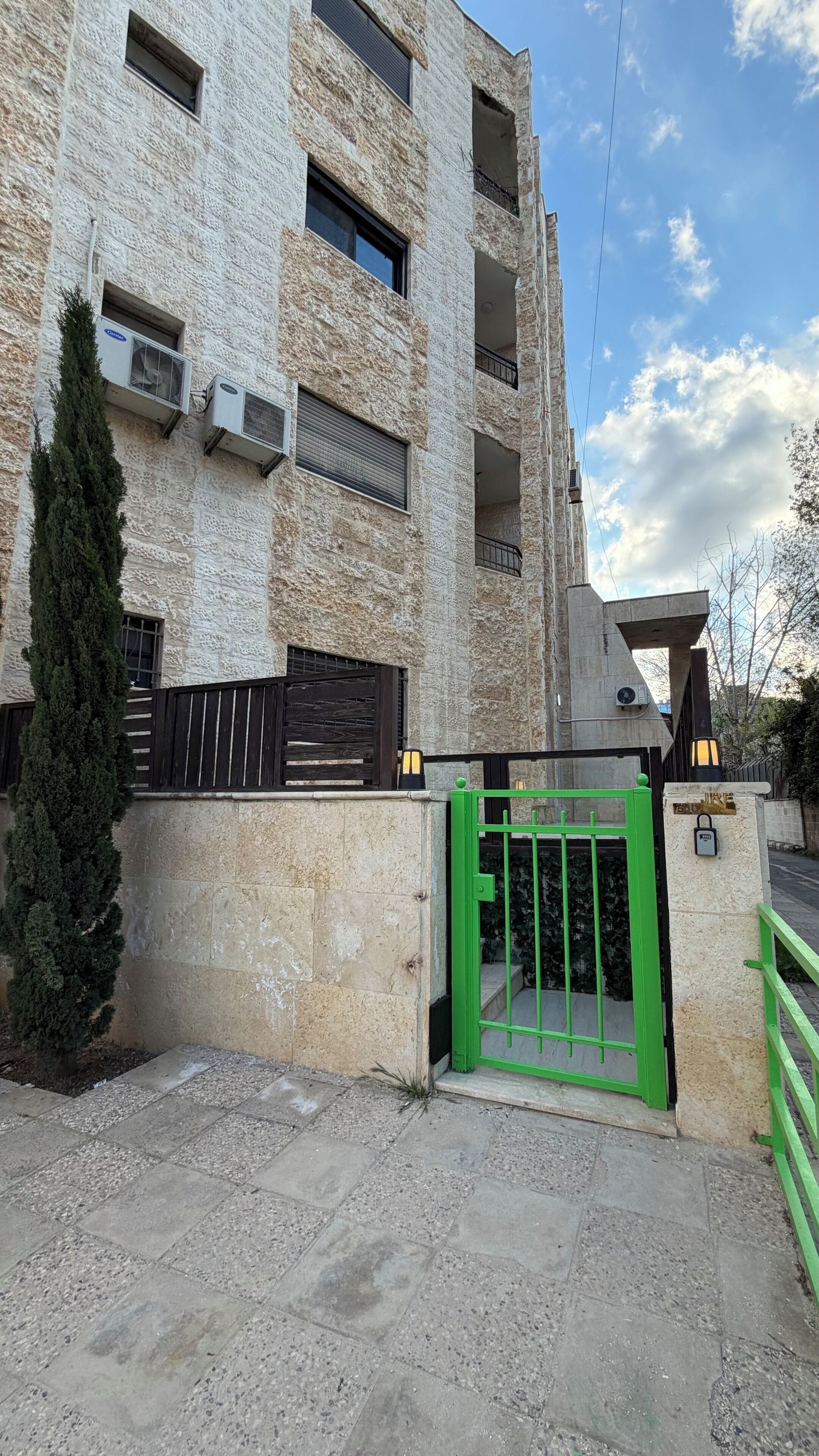 A multi-story building with a stone facade, air conditioning units, a green gate, and a small tree.
