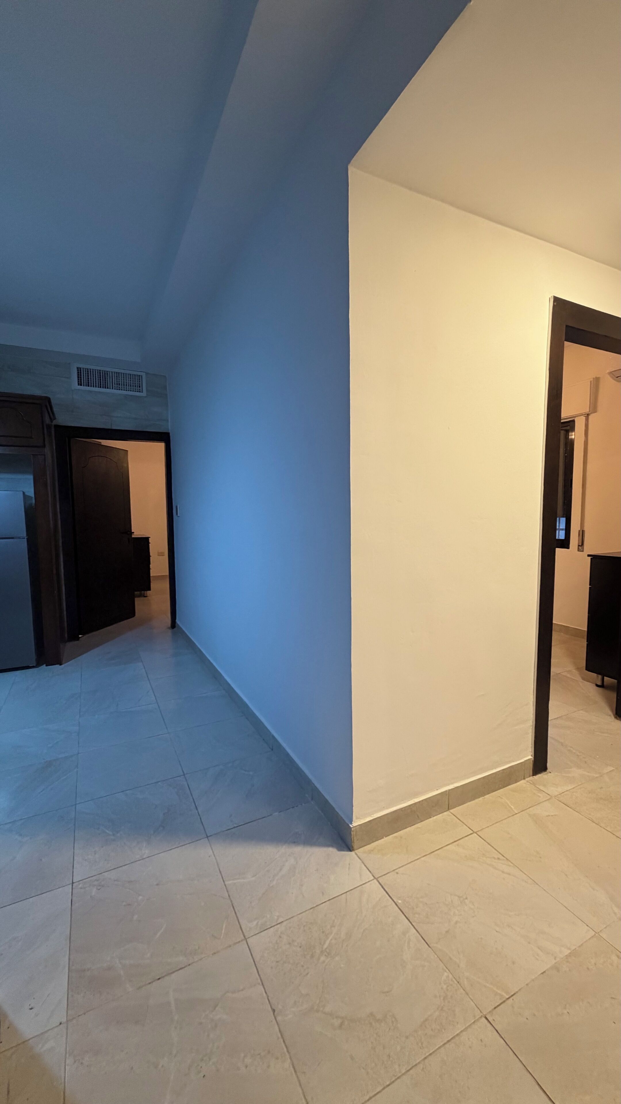 Un pasillo con suelo de baldosas, una puerta a la derecha y una rejilla de ventilación en la pared izquierda.