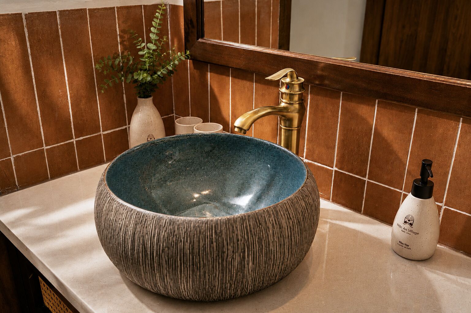 Ein Badezimmer mit einem runden, strukturierten blauen Waschbecken, einem goldenen Wasserhahn, einer weißen Vase mit Grünpflanzen und einer Handseifenflasche.