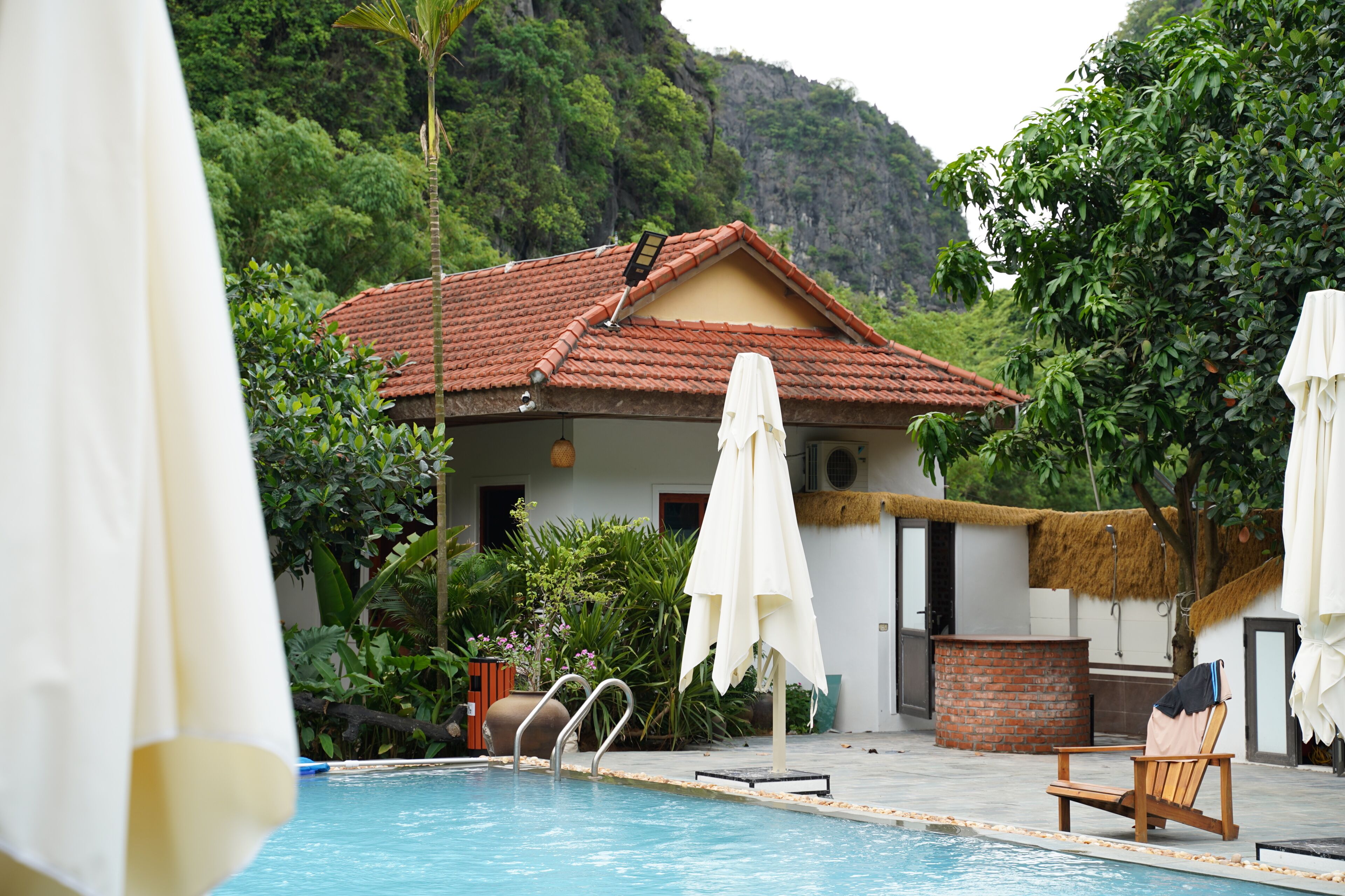 A pool area with a red-tiled roof building, white umbrellas, and a wooden chair.