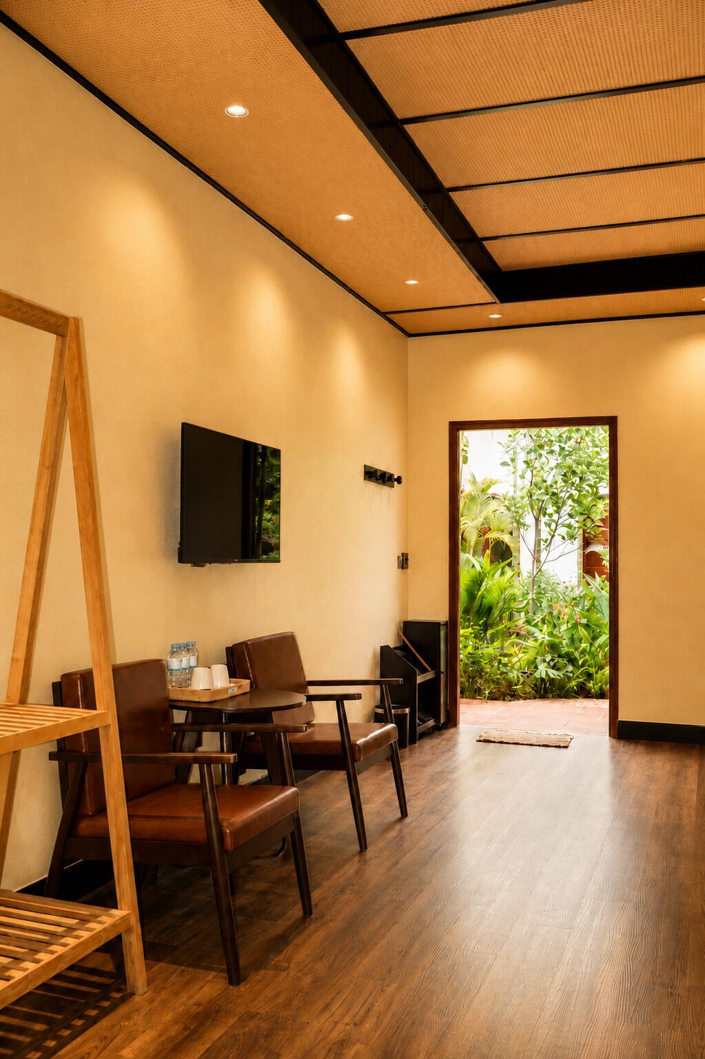 A modern hotel room with a wooden floor, a dining area with chairs and a table, a mounted TV, and a view of greenery outside.