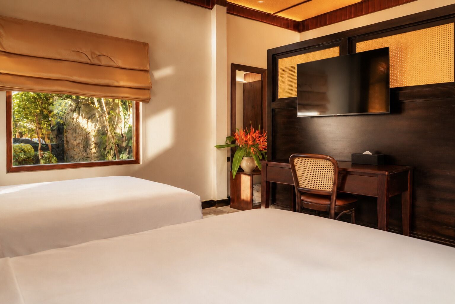 A hotel room with a bed, a desk, a chair, a TV, and a window with a view of greenery.