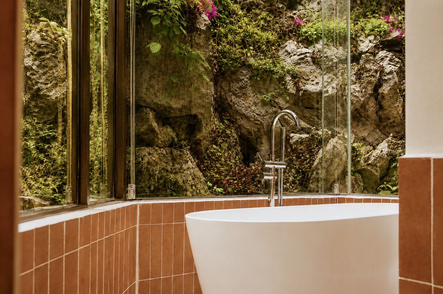 A bathroom with a large bathtub, a glass shower enclosure, and a view of a rocky wall with vegetation.