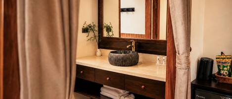 Un baño con tocador de madera, un lavabo redondo, un espejo, un perchero para toallas y una máquina de café.