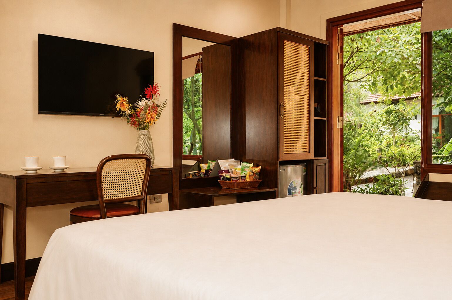 A hotel room with a bed, a desk with a chair, a television, a window with a view of greenery, and a small refrigerator.