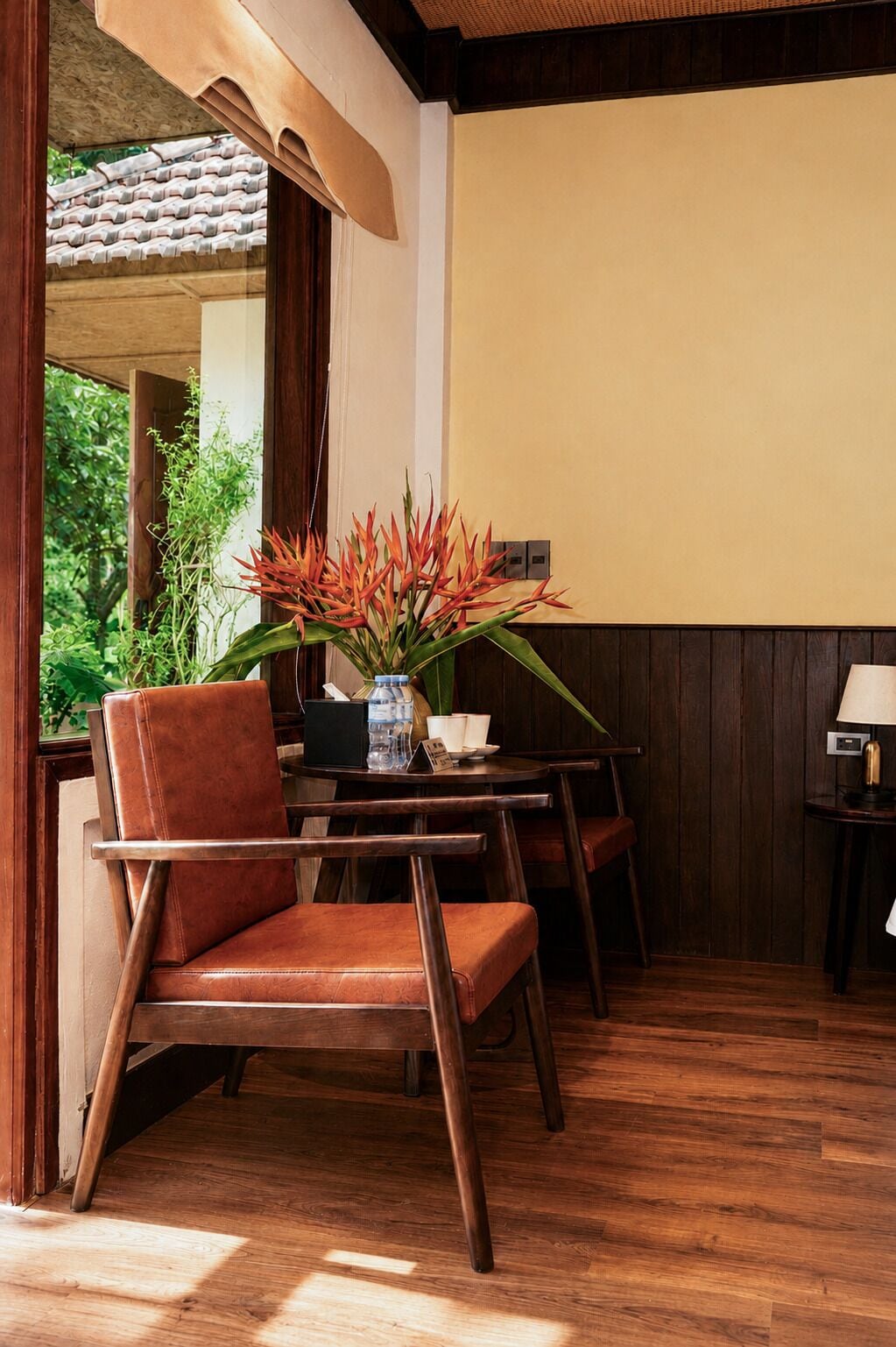 An indoor seating area with wooden furniture, a table with a vase of flowers, and a view of greenery outside.