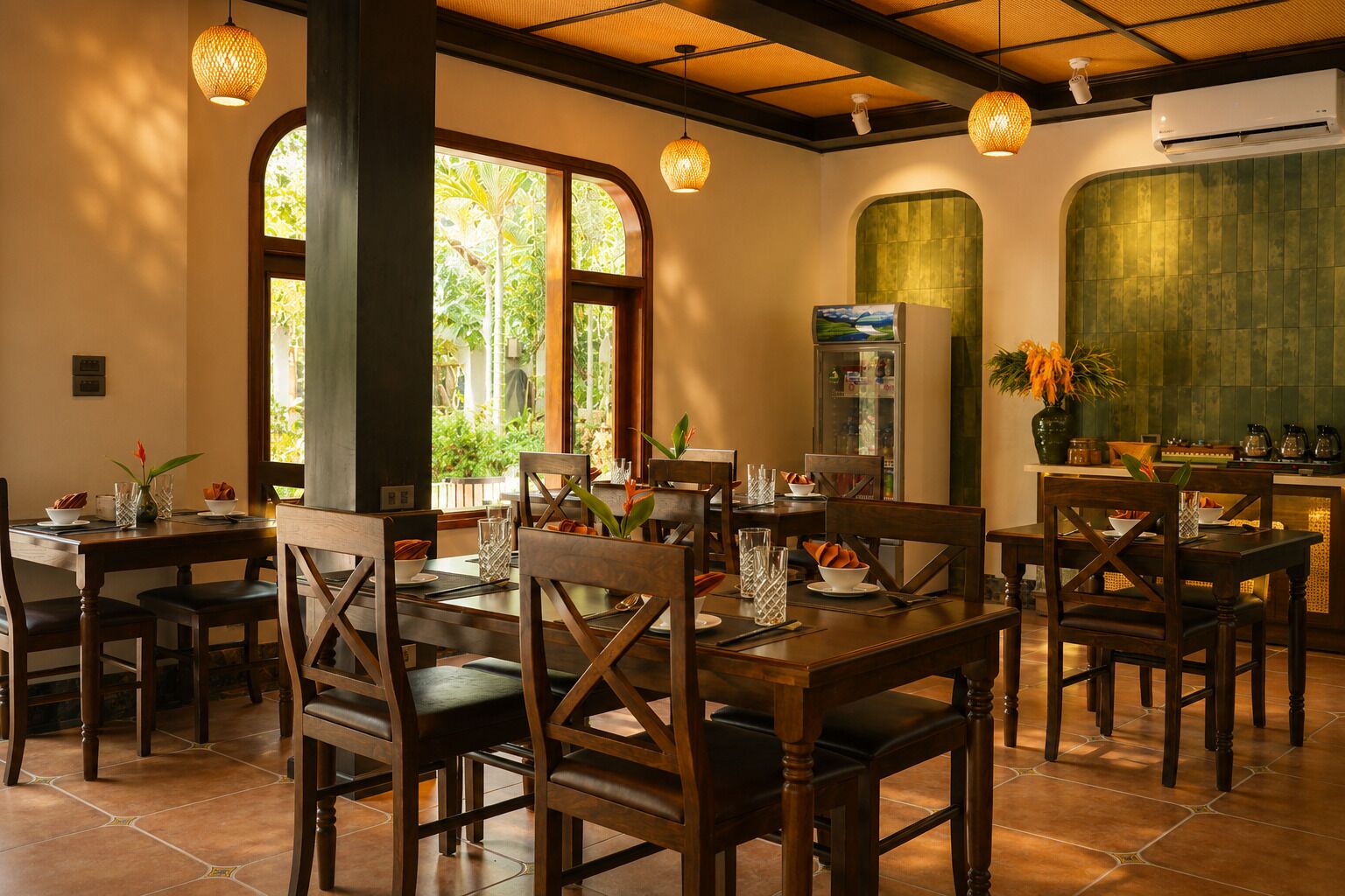A restaurant with wooden tables and chairs, large windows with greenery outside, and hanging light fixtures.