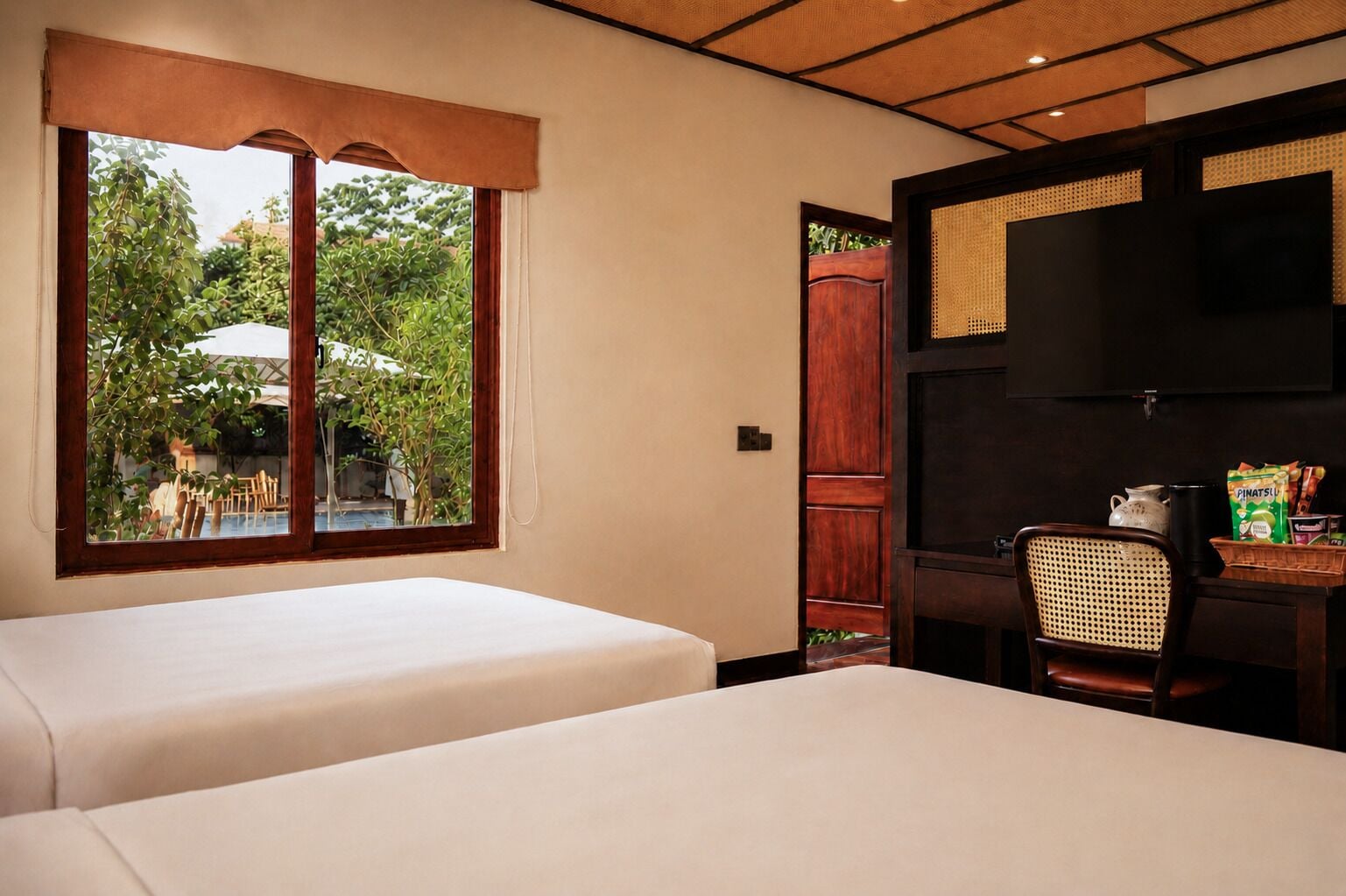A hotel room with two beds, a window with a view of greenery, a wooden door, and a small table with a chair.