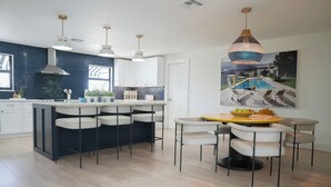 Una cocina moderna con una isla central, taburetes de bar, iluminación colgante y un comedor con una mesa redonda y sillas.