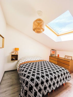 Une chambre avec un puits de lumière, un lit avec une literie à motifs, une commode en bois et une petite table de chevet avec une lampe.