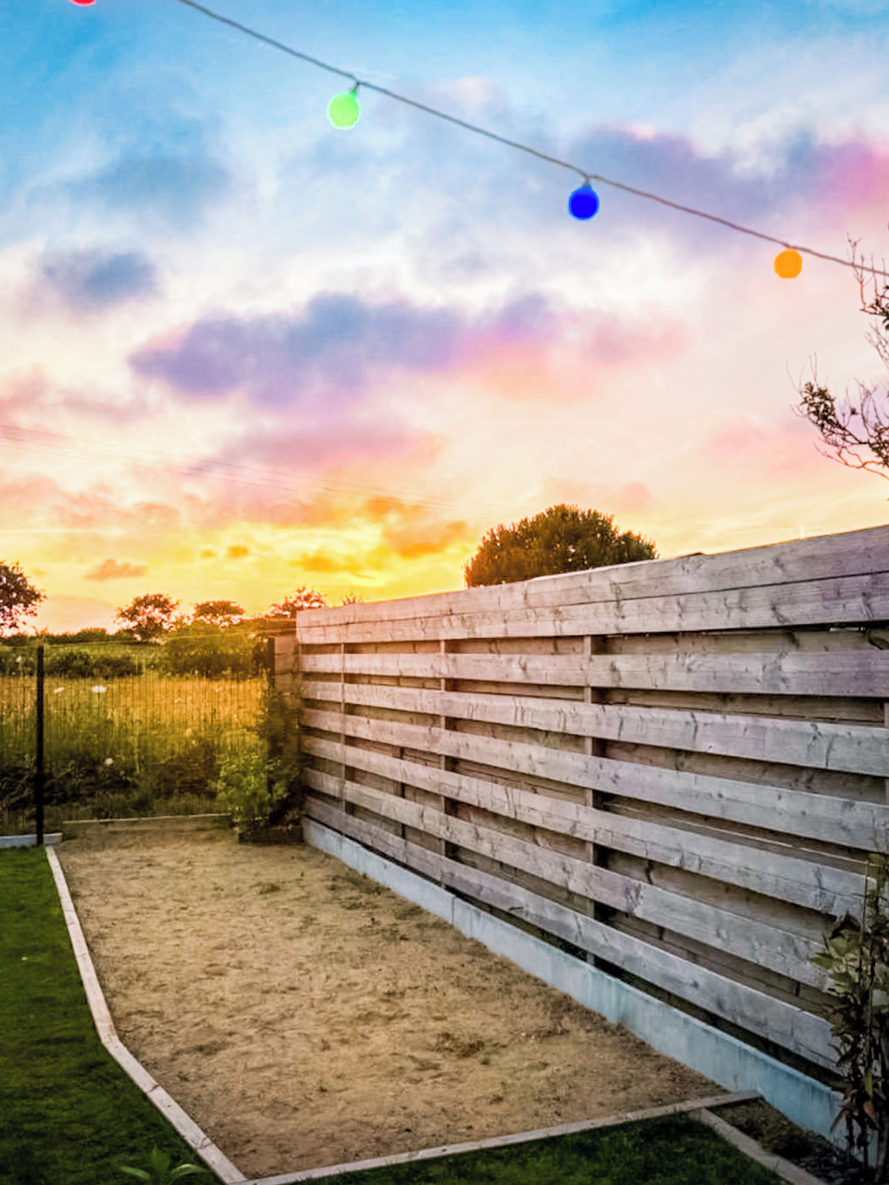 A wooden fence, a grassy area, and a string of lights against a sunset sky.