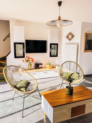 Un salon moderne doté d’un téléviseur à écran plat, de fauteuils en osier, d’une table basse en bois et d’un luminaire suspendu au plafond.