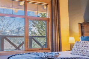 A room with a large window, a bed with a patterned pillow, a lamp, and a wooden headboard. - Hygge House - downtown mountain view sanctuary (Canmore)