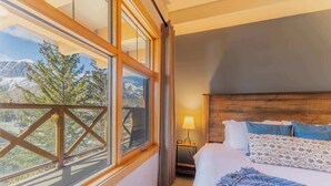 Ein Hotelzimmer mit großem Fenster, einem Bett mit Kissen, einem Nachttisch mit Lampe und Blick auf die Berge.