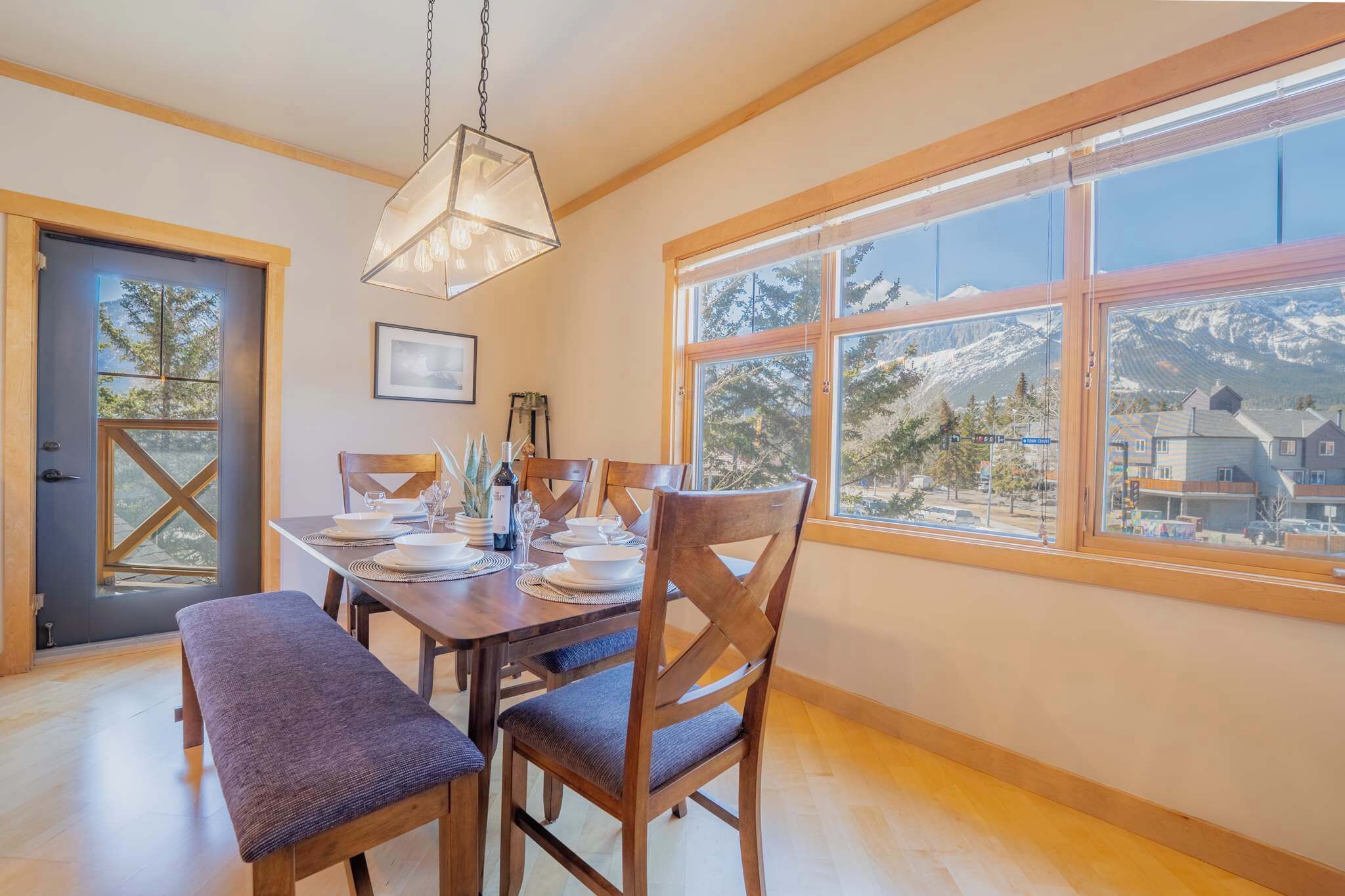 Comedor con una mesa lista para cuatro personas, sillas de madera, un banco y un gran ventanal con vistas a la montaña.