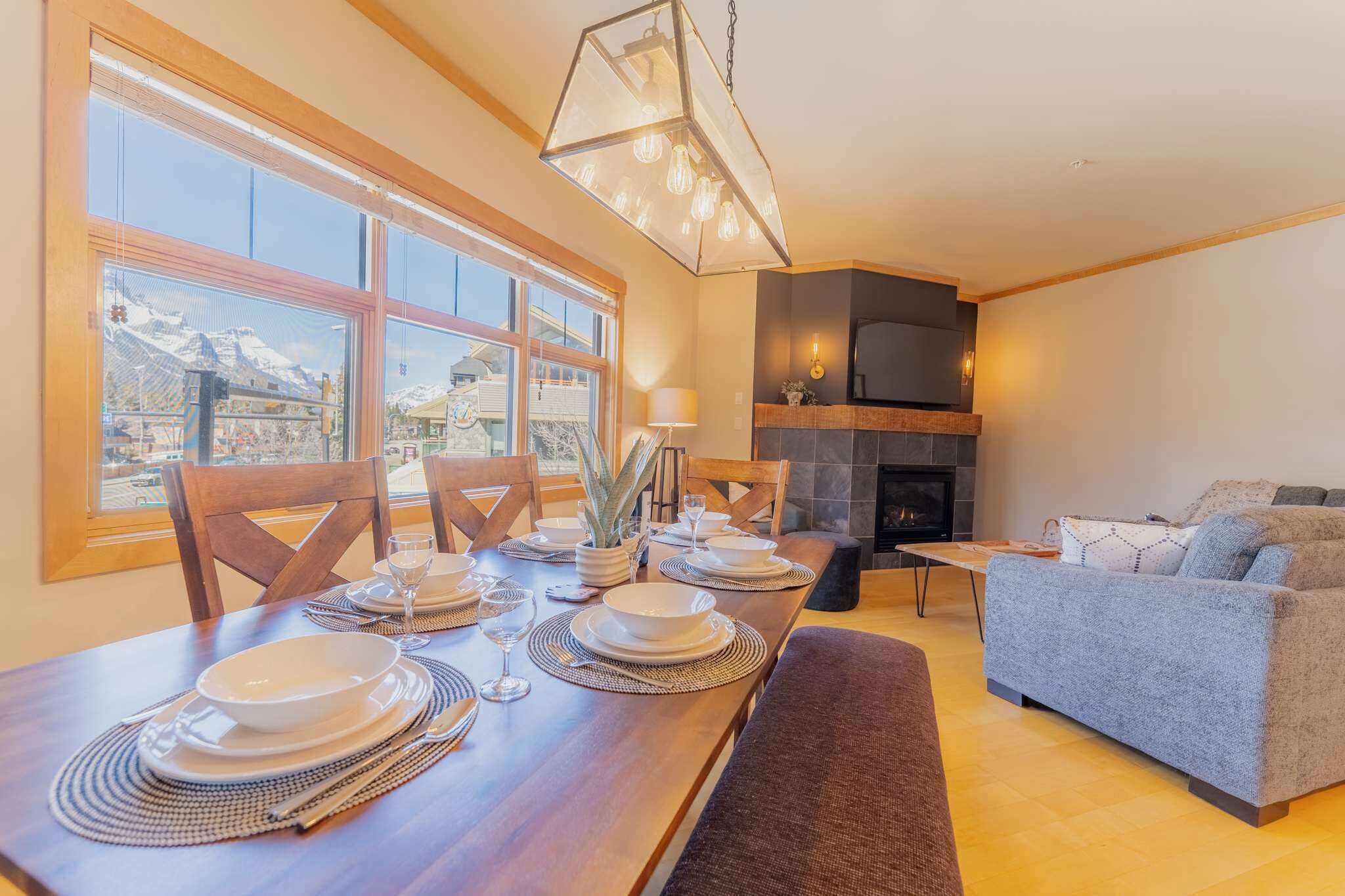 A dining area with a wooden table set for four, a sofa, a fireplace, and a view of mountains through large windows.