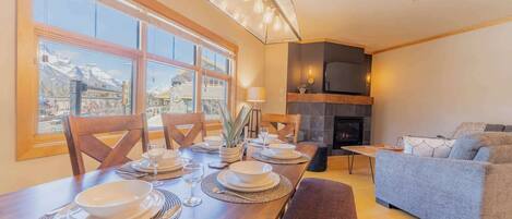 A dining area with a wooden table set for four, a sofa, a fireplace, and a view of mountains through large windows.