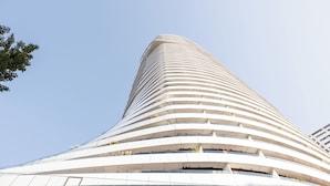 A modern, curved skyscraper with multiple balconies and a clear sky in the background. - Skyline Suite - Danube-View Balcony&Pool (Vienna)