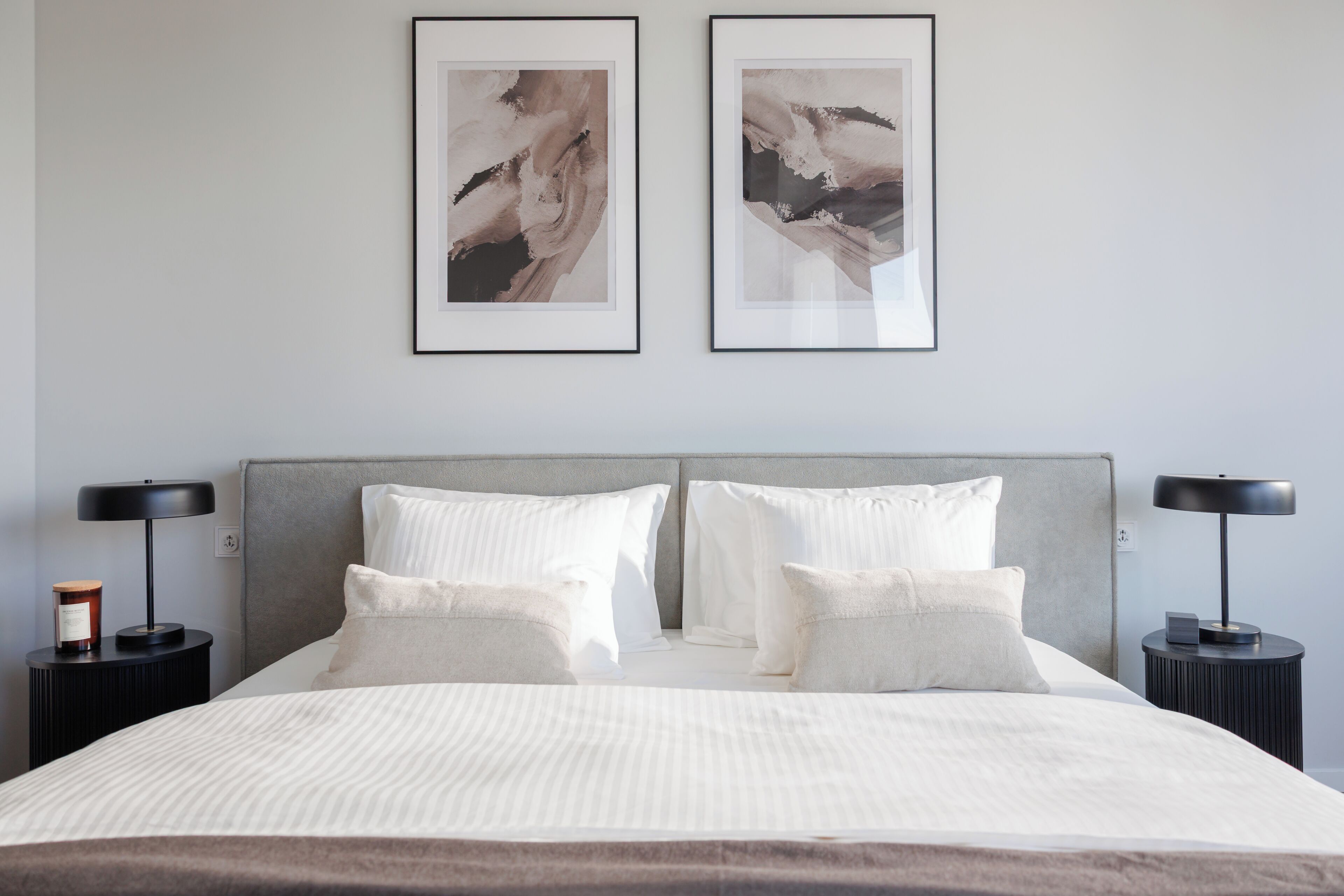 Un lit bien fait, avec du linge de lit blanc, deux tableaux abstraits encadrés accrochés au mur et deux tables de chevet avec des lampes.