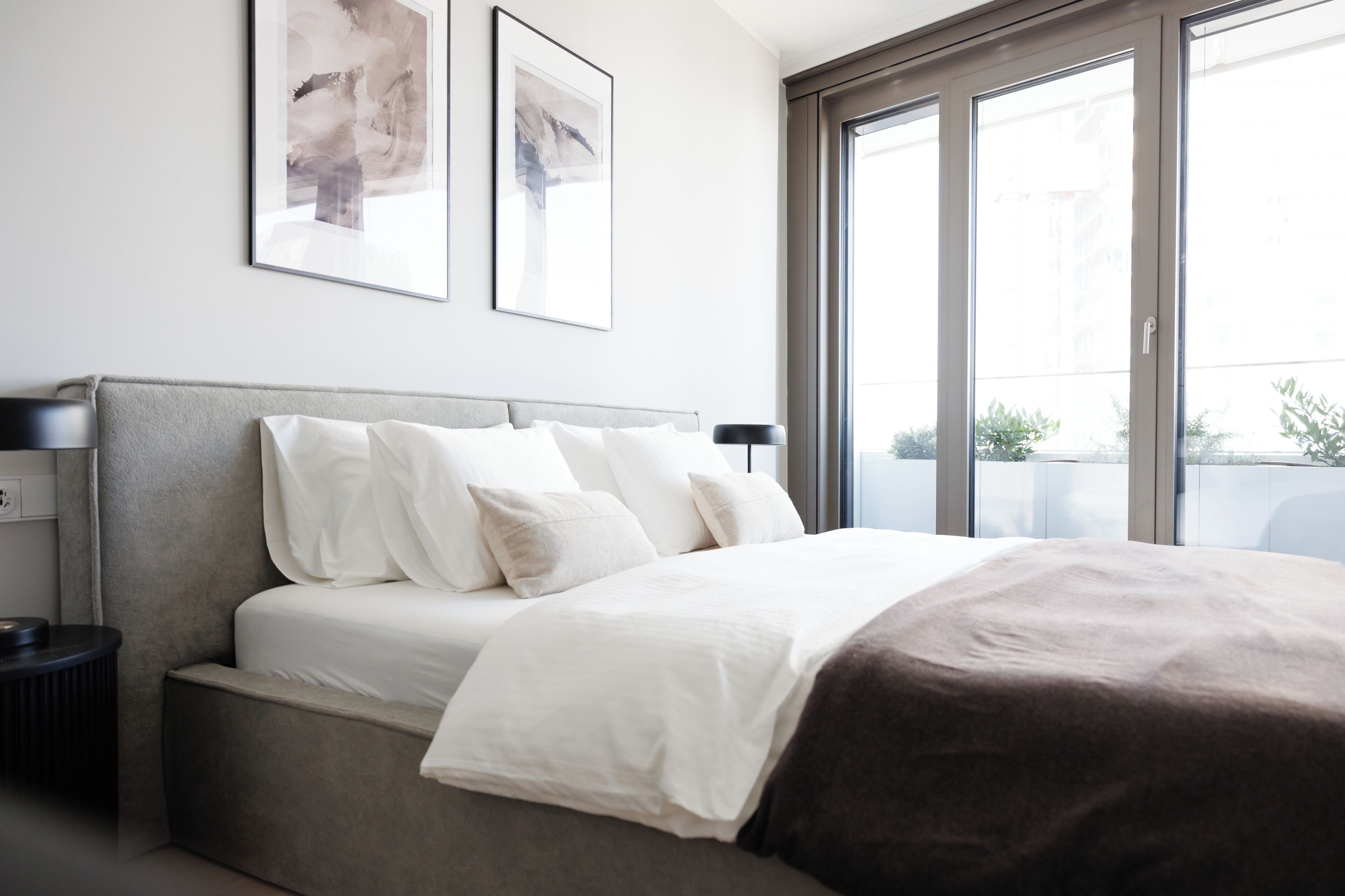 A modern bedroom with a bed, a nightstand, a lamp, and large windows.