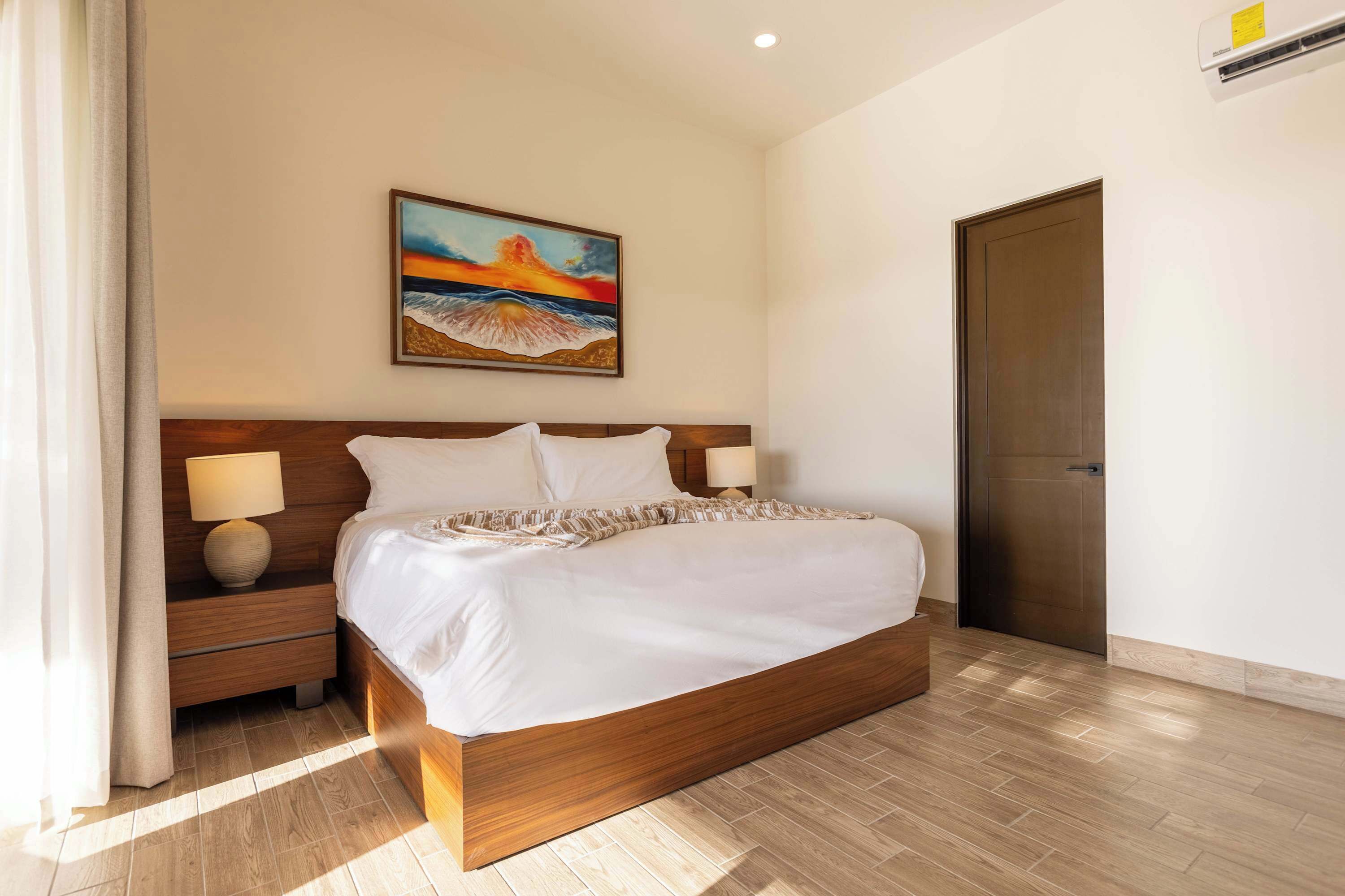 A hotel room with a large bed, two bedside tables with lamps, a painting of a sunset, and a door.