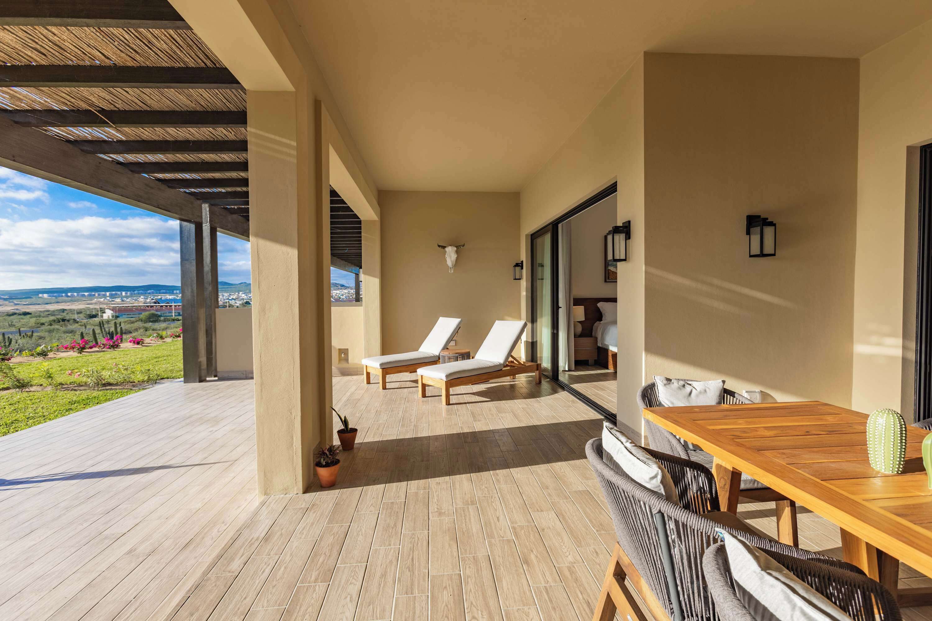 A covered outdoor area with a wooden table and chairs, a pair of sun loungers, and a view of the landscape.