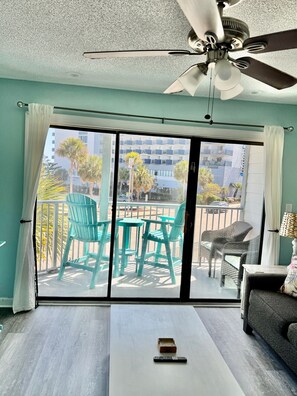 A room with a ceiling fan, a sliding glass door leading to an outdoor area with chairs, a sofa, and a table with a remote control. - Incredible Beach Villa 1bd/1ba Myrtle Beach (Myrtle Beach)