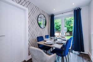 A dining area with a table set for four, a mirror on the wall, and a view of greenery outside. - House Sleeps 7 With Garden and Free Parking (Manchester)