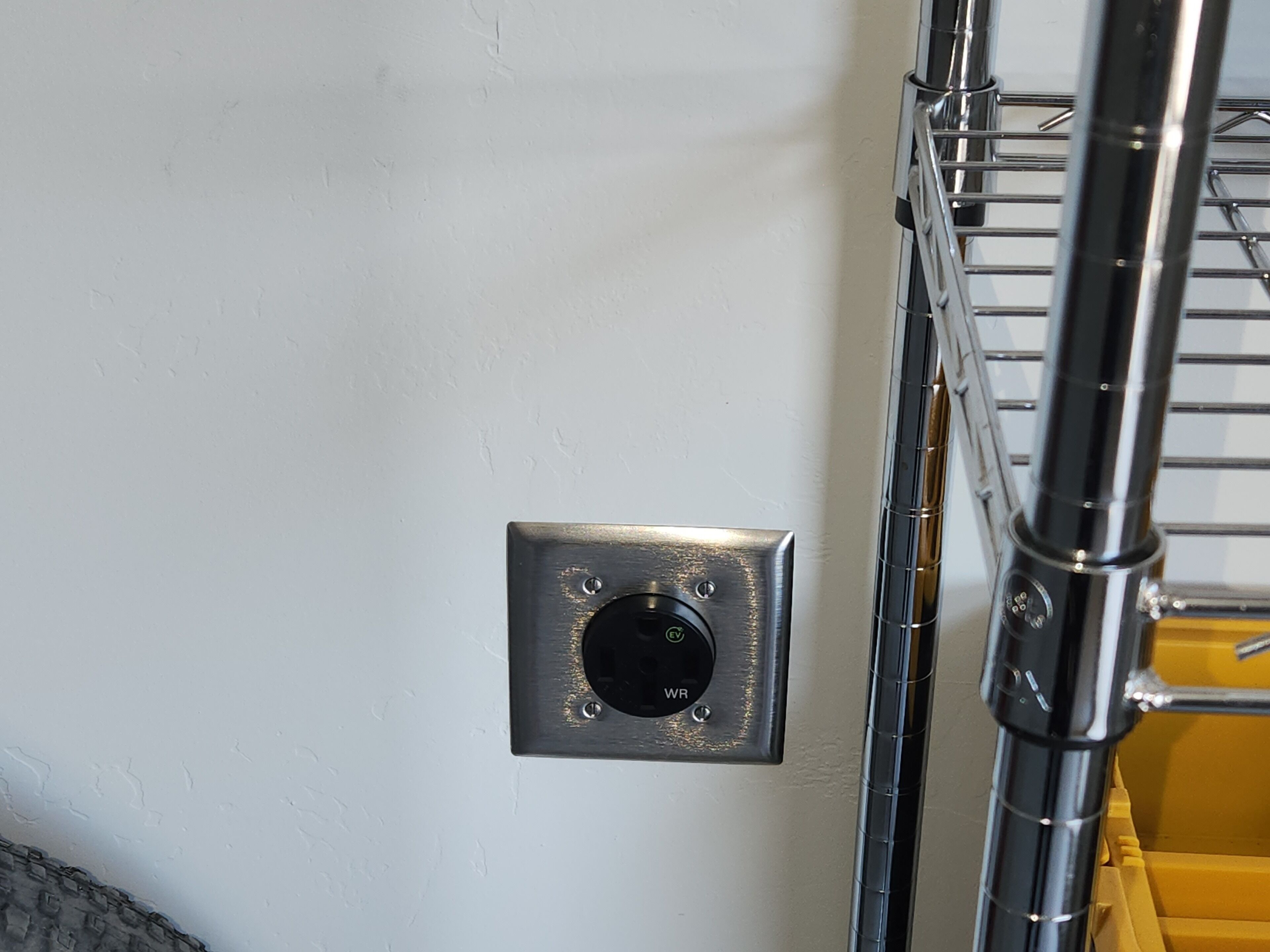 Un enchufe eléctrico metálico en una pared blanca, una repisa metálica con un recipiente amarillo y una estructura metálica con una abertura circular.