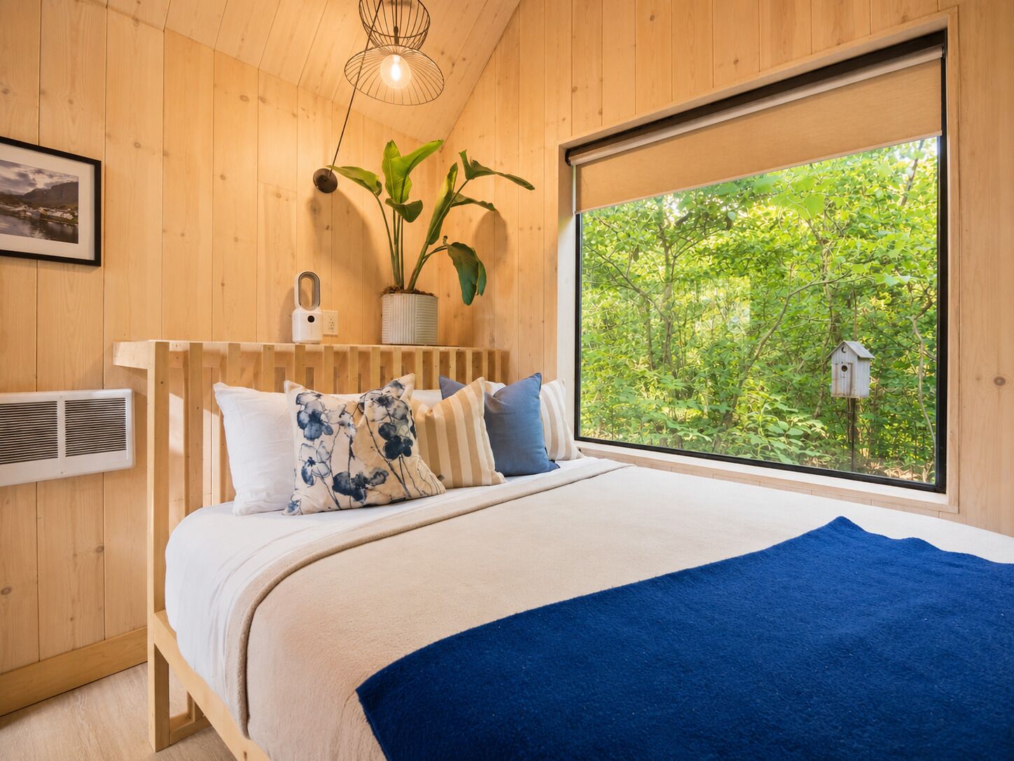 Une chambre avec une grande fenêtre, un lit recouvert d’une couverture bleue, une plante en pot et un luminaire fixé au mur.