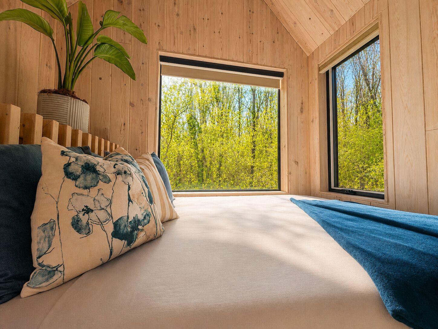 Un dormitorio con un ventanal grande, una cama con manta azul y una planta en maceta.
