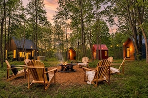 A forest setting with a fire pit and wooden chairs arranged for outdoor seating.