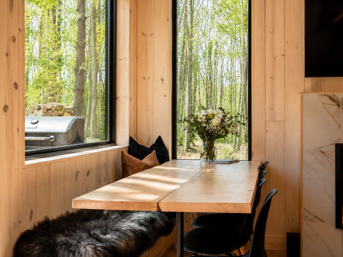 A wooden interior with a dining table, chairs, a fur rug, a fireplace, and a window with a view of trees.