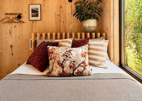 A wooden room with a bed, pillows, a hanging plant, and a window with a view of greenery.