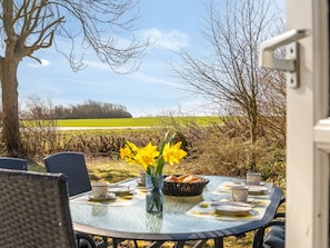 Et udendørs spiseområde med et bord dækket til fire personer, omgivet af stole, en vase med blomster og en kurv med brød.