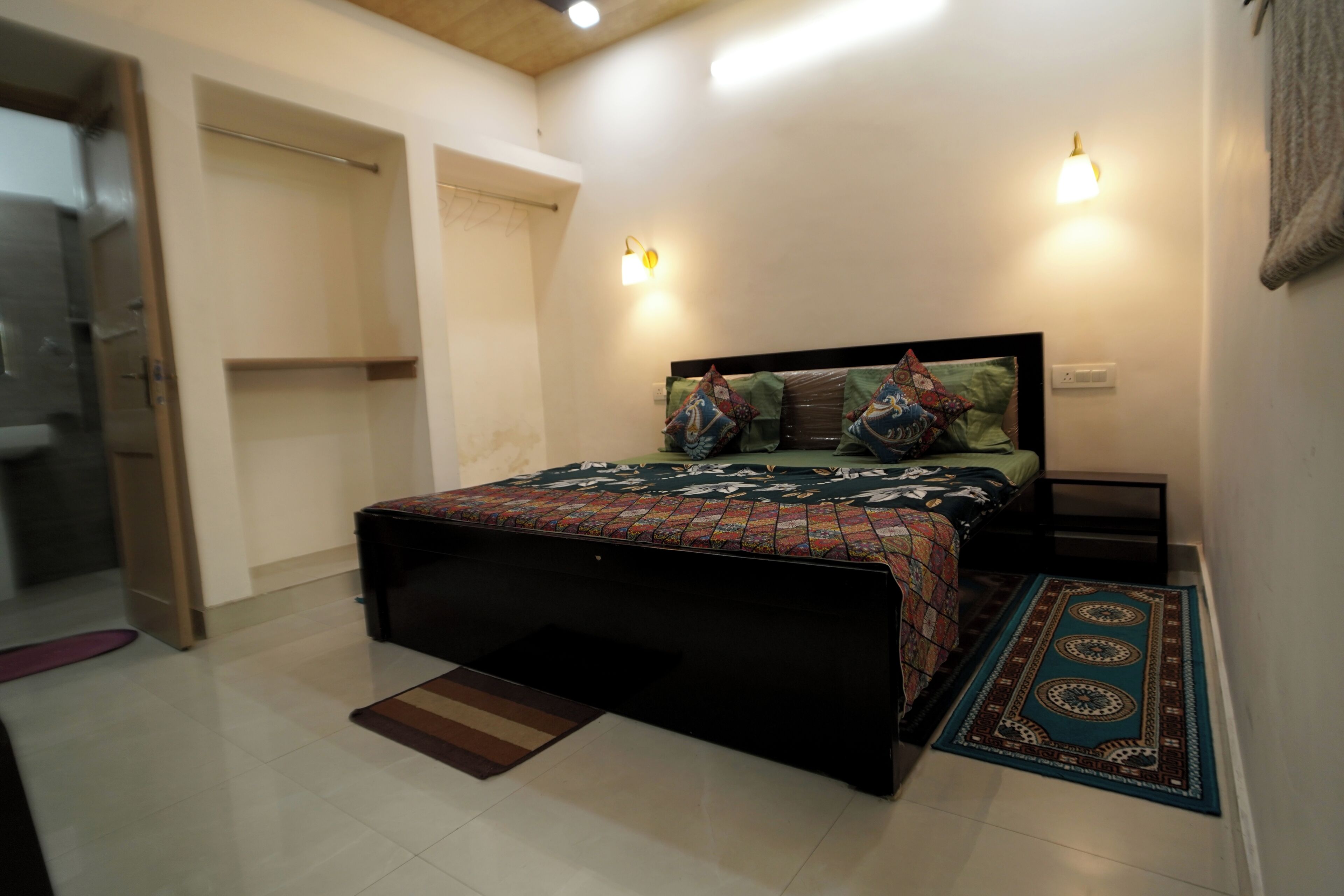 Un dormitorio con cama, mesita de noche, lámpara de pared, puerta y alfombra estampada.