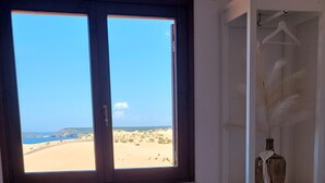 Quarto com uma grande janela com vista para uma praia, cama com toalhas brancas dobradas e um guarda-roupa branco com cabideiro.