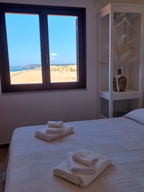 A bedroom with a large window overlooking a beach, a bed with neatly folded white towels, and a white wardrobe with a hanging rack. - Dune nel Blu (Torre dei Corsari)