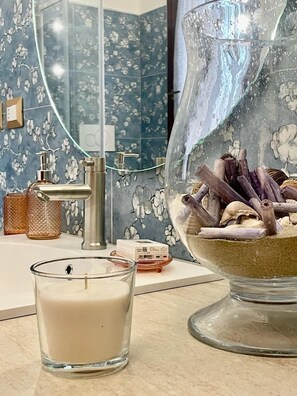 Ein Badezimmer mit blumigem Fliesenbild, einem modernen Wasserhahn, einem Glas-Teelicht-Halter mit Dekoration und einem Teelicht auf der Ablagefläche.